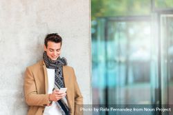 Smiling man leaning on wall outside texting and wearing a camel coat  47mxz6