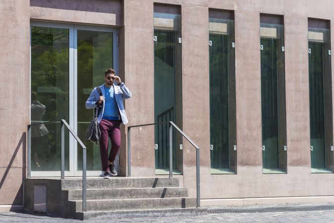 Businessman leaving office building while holding a shoulder bag outdoors adjusting sunglasses
