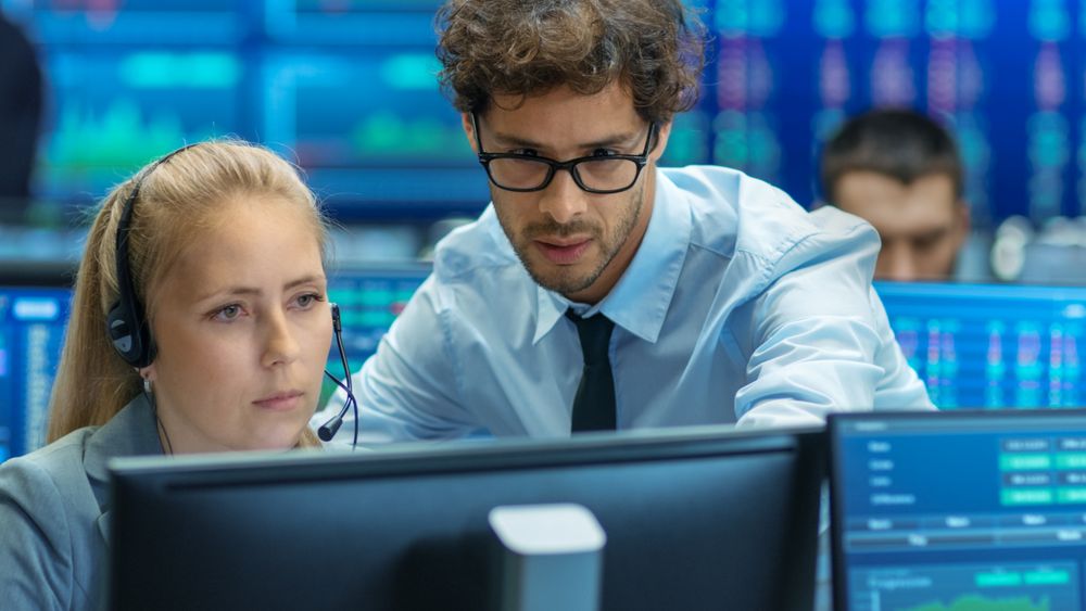 Experienced Stock Exchange Trader Teaches His Apprentice at Her Workstation.