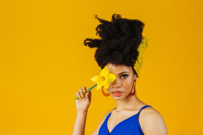 Portrait of Black woman with large earrings holding a daffodil over her eye