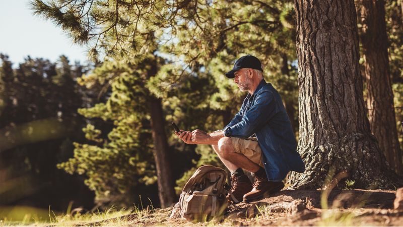 Mature man hiker sitting by a tree in forest and checking his position by using a compass
