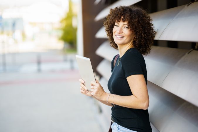 A cheerful woman with curly hair joyfully holding a tablet while enjoying the outdoors