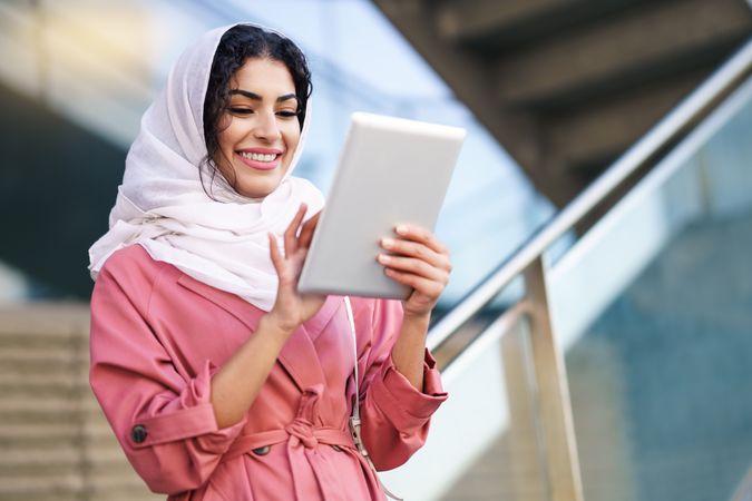 Smiling female in headscarf and pink trench coat using her digital tablet