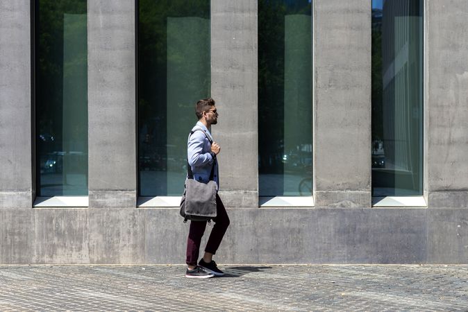 Side view of man walking in the sun outside of building with shoulder bag