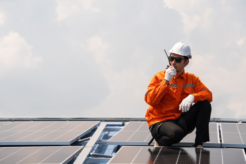 Male engineer in protective work wear talking on walkie talkie with colleague while checking on solar cell or photovoltaic panel installed on the roof of the factory. Industrial renewable energy