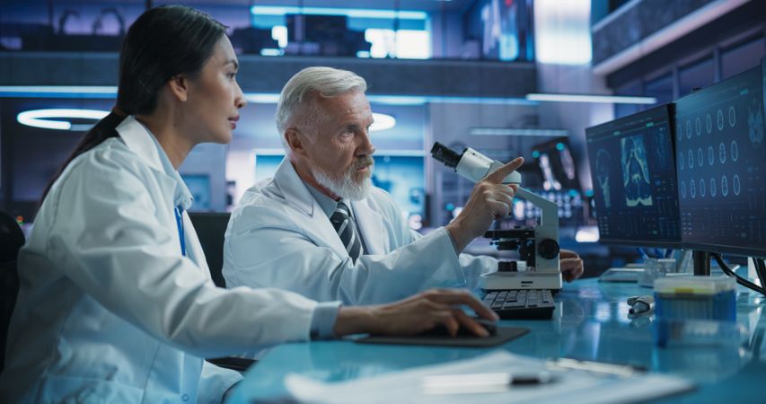 Biotechnology Medical Research Center: Female Asian Neurologist Using Desktop Computer And Talking To Male Scientist With Microscope.