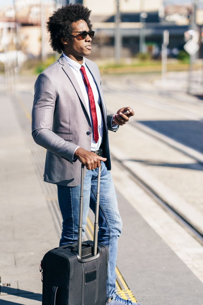 Man in jeans and dress shirt with tie standing outside with roller suitcase