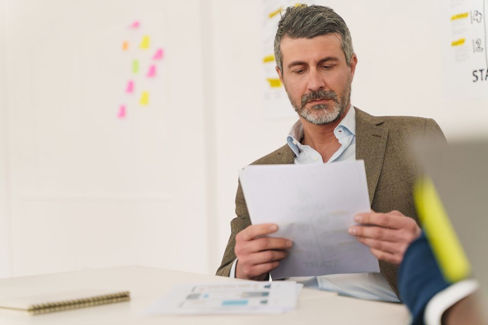 Grey haired businessman with  beard, sitting at desk, reading a document