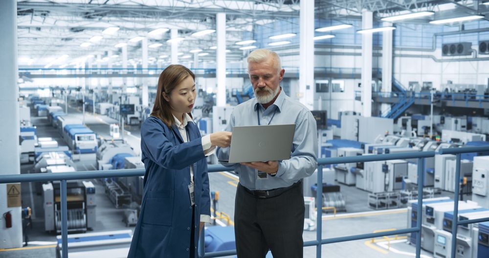 Male Engineer and Female Project Manager Standing on a Platform, Talking, Using Laptop Computer and Monitoring Production at a Modern Automated Electronics Manufacture with AI Robots