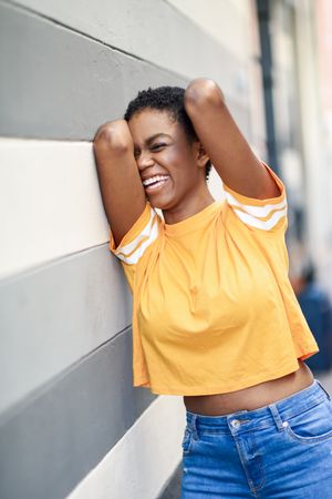 Black woman leaning on grey striped wall and smiling with arms up, vertical composition