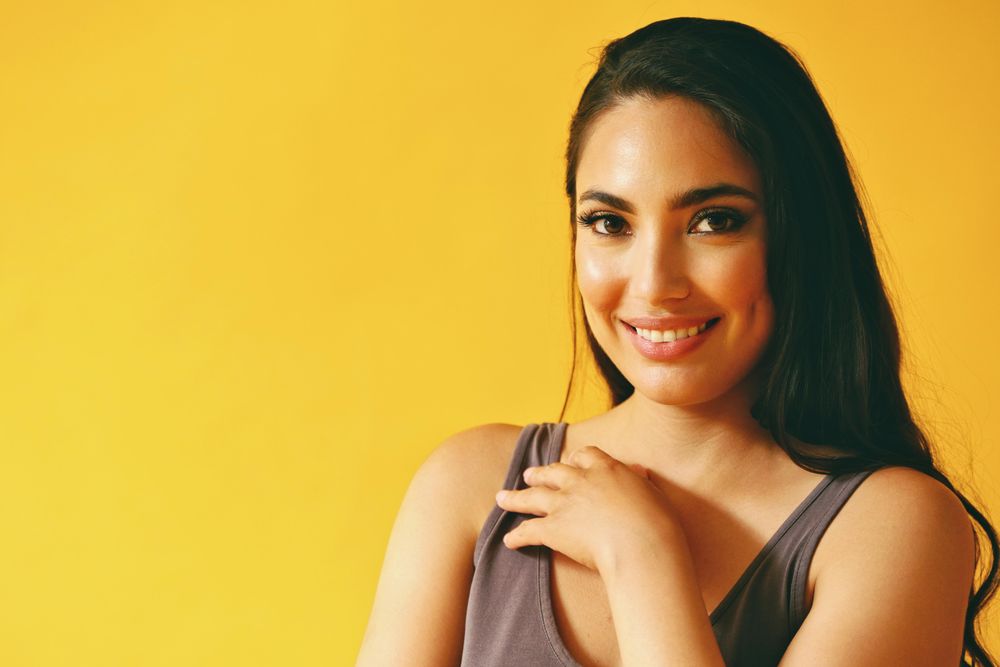 Happy confident Hispanic woman looking at camera in yellow room with her hand to her chest