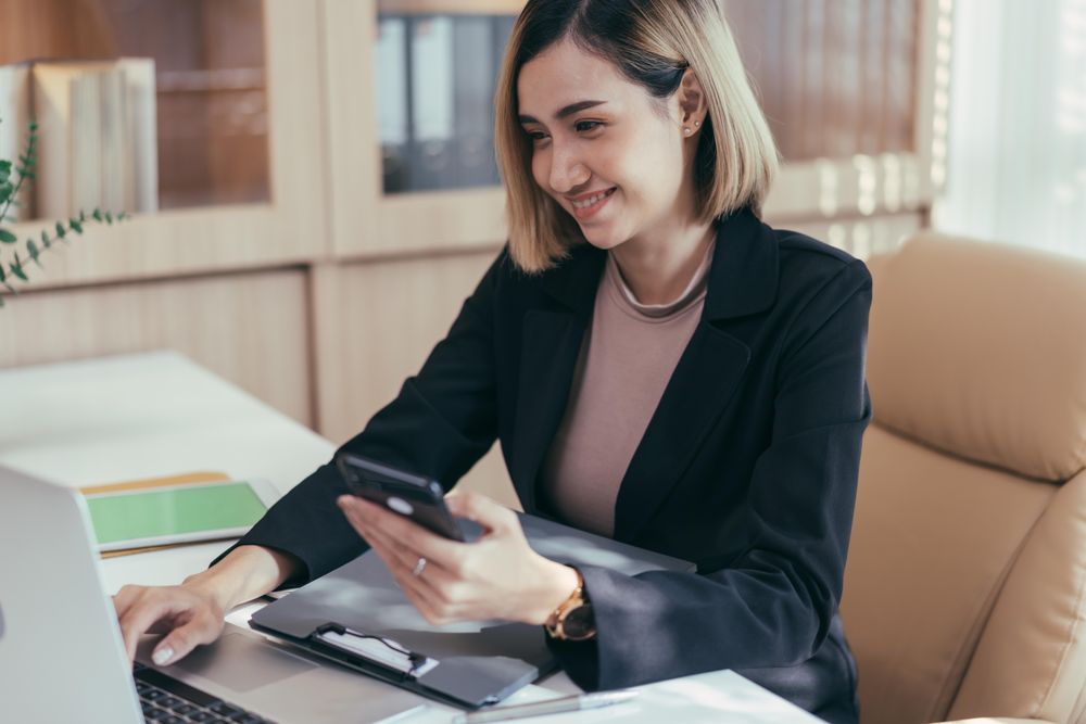 Woman smiling at phone while sitting in her office with laptop