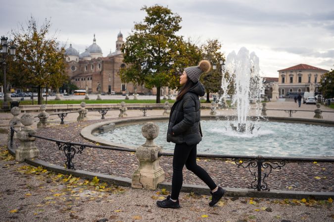 Young female tourist in warm wear strolling at ancient park with beautiful buildings and fountain