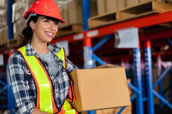Smiling female warehouse worker in safety gear holding a box.