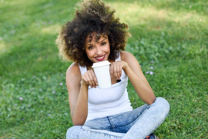 Black woman with afro hairstyle sitting cross legged in park sipping coffee