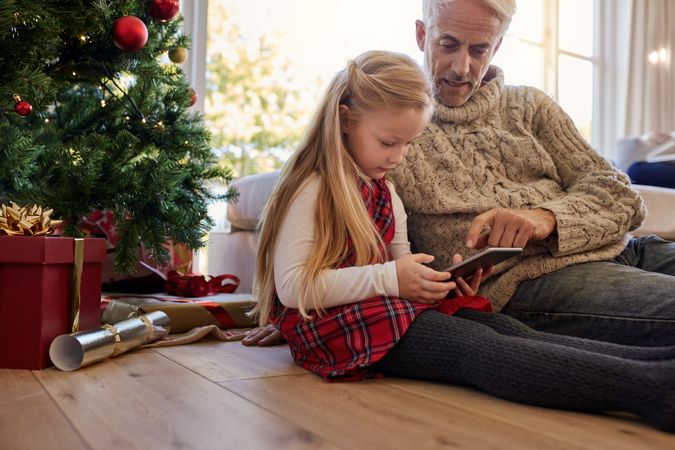 Little girl with grandfather sitting by christmas tree and using digital tablet