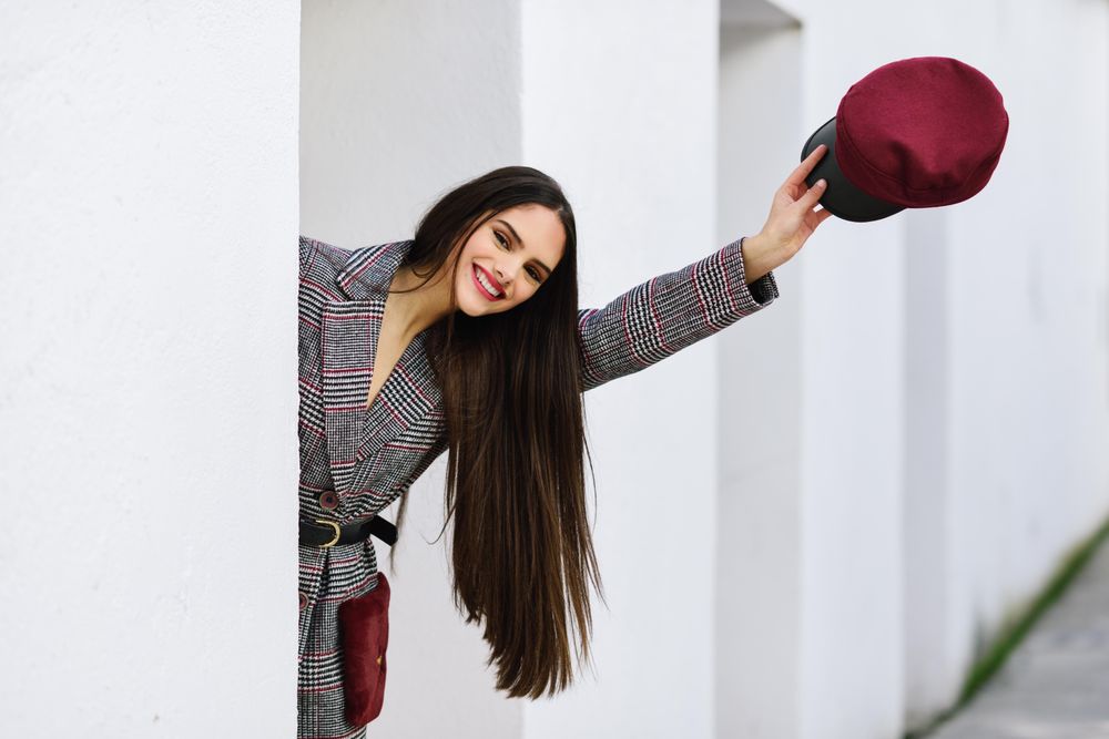Smiling female peaking out from behind light wall outdoors holding out her red cap