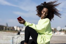 Carefree smiling female with curly hair in green shirt sitting listening to music on pier