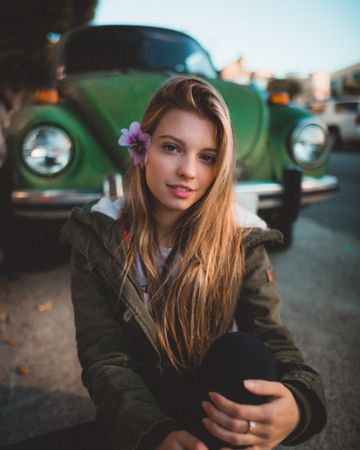 Woman in green jacket smiling and sitting on ground near green Volkswagen car