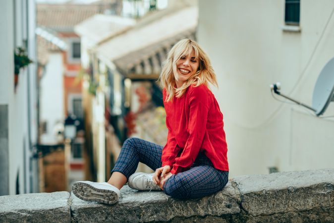 Happy blond woman sitting on stone wall with legs crossed above Spanish street