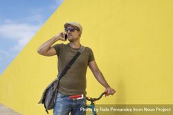 Man in hat and sunglasses talking on phone standing with bike next to yellow wall 0LmWX5