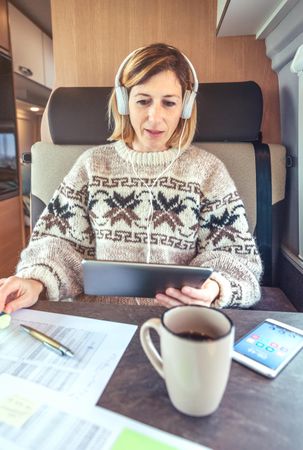 Female sitting in back of camper van working on digital tablet with headphones, vertical