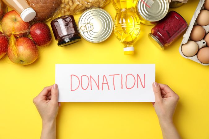 Female hands hold text Donate and different food on yellow background, top view