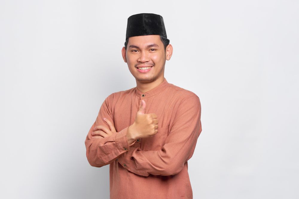 Happy Muslim man in kufi hat making thumbs up gesture with hand
