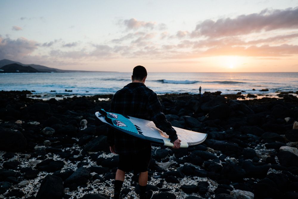 Back of man with surfboard walking towards ocean on rocky terrain