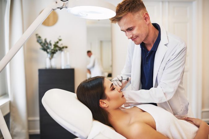 Smiling male dermatologist injecting treatment into brunette patient’s face