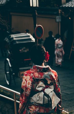 Back view of Japanese woman in red kimono walking near rickshaw on street in Osaka, Japan