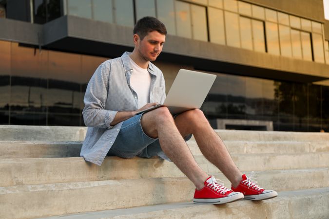 Young man sits in steps and working in laptop outdoor. Freelancer