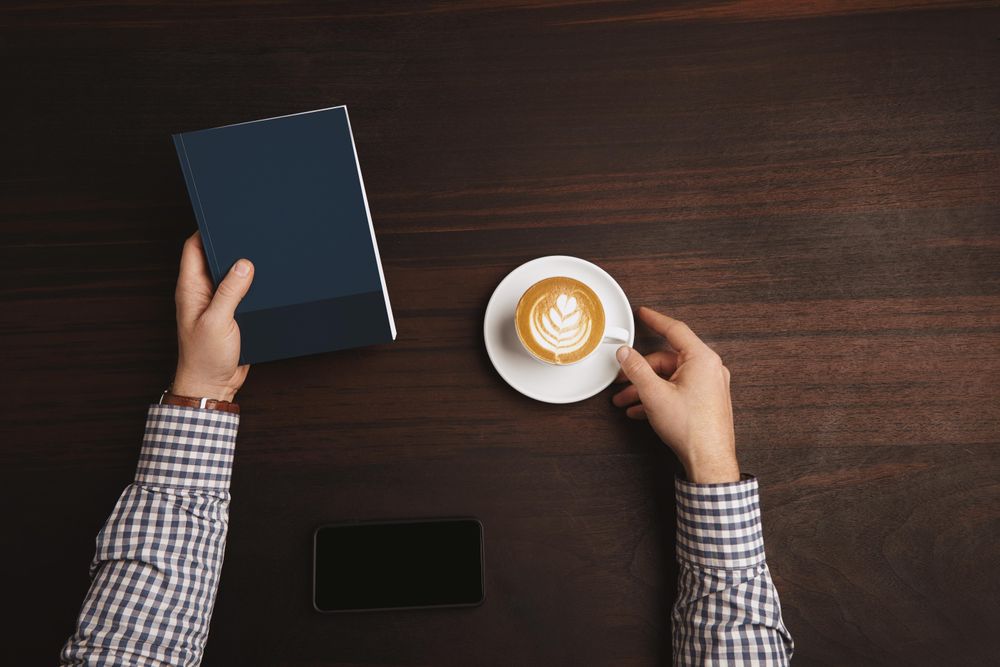 Arms in plaid shirt and leather wrist watch with cappuccino and closed notebook