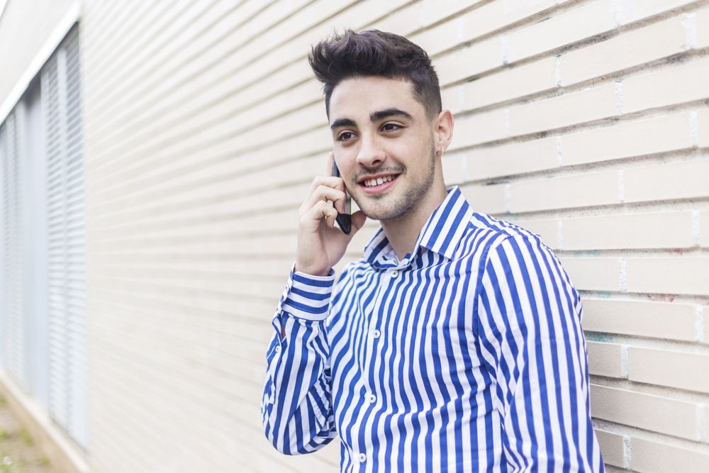 Young man leaning on house wall talking on his smartphone and looking into the distance