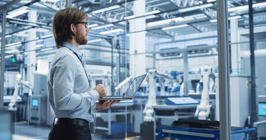 Factory Manager Working on Laptop at a Futuristic Industrial Manufacture with Autonomous Support Robots. Specialist Monitoring Technical Data on a Computer, Analyzing Reports and Planning Updates