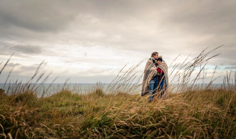 Couple cuddling while wrapped in blanket on a windy day in the long grass above the coast
