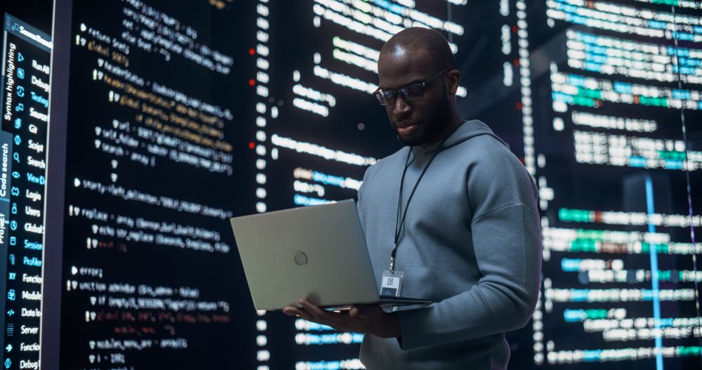 Portrait of Young Black Man Working on Laptop, Looking at Big Digital Screen Displaying Back-end Code Lines.