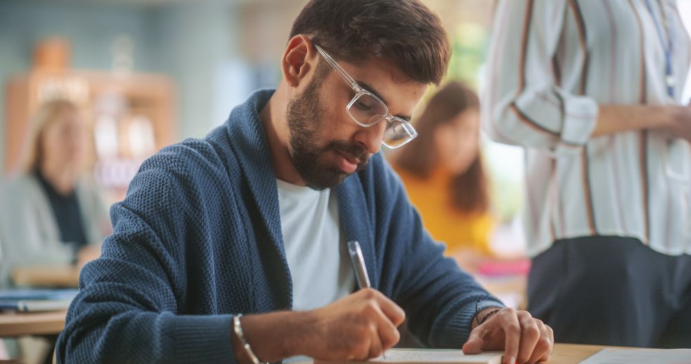 Young Man Wearing Glasses, Sitting Behind a Desk and Writing Down Notes Together with Diverse Colleagues