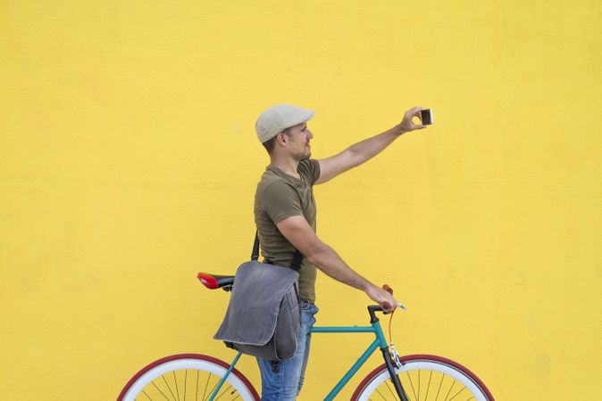 Male in hat and sunglasses taking photo on phone while sitting on colorful bicycle
