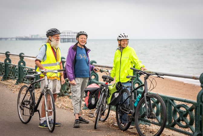 Three smiling older people standing with bikes looking away by coast