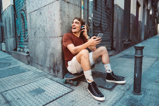 Relaxed man in headphones using smartphone while resting on longboard on street
