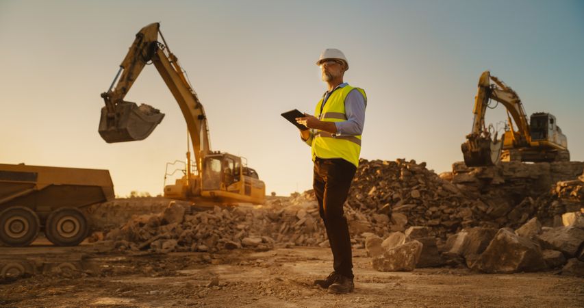Male Real Estate Investor Wearing Protective Goggles And Using Tablet On Construction Site On Sunny Day. Man Inspecting Building Progress. Excavator Loading Materials Into Industrial Truck