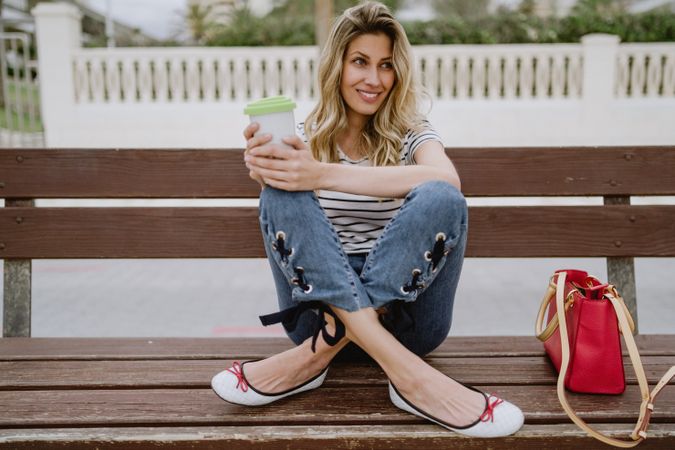 Happy woman with disposable cup of coffee sitting on street bench