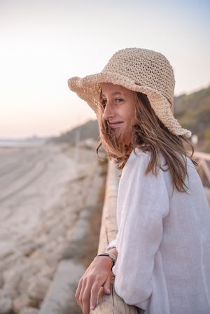 Charming woman in hat leaning on fence and looking at sea