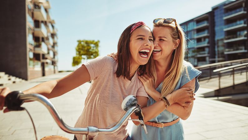 Two laughing female friends with arms on each others shoulders while walking with bike, copy space