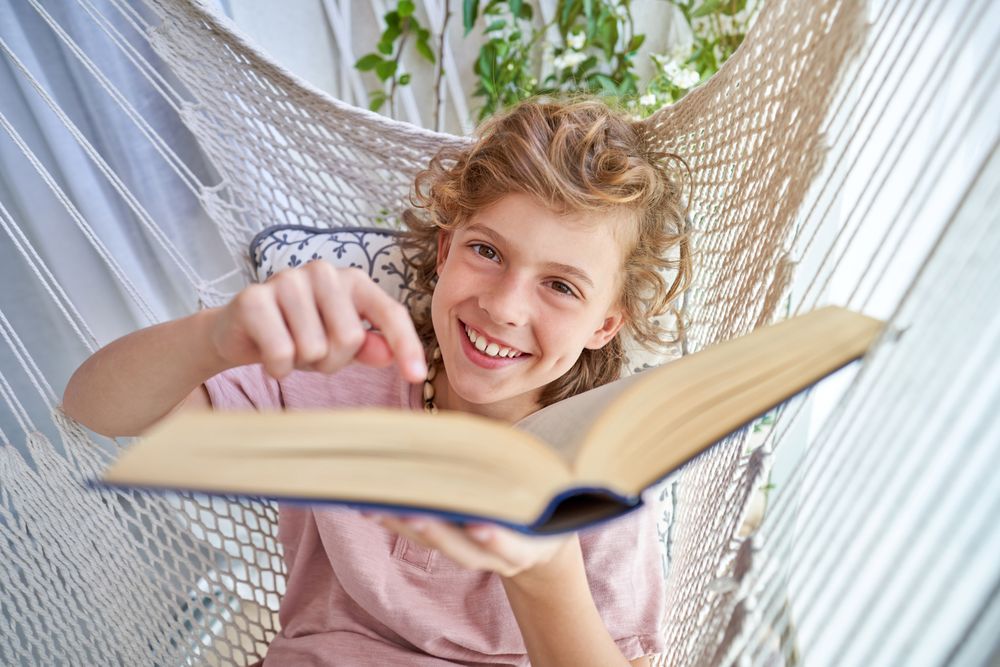 Positive boy lying in hammock and pointing at book