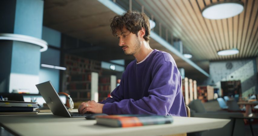 Student Studying in a Modern Library. Young Man Thinking and Problem Solving School Exercises. Male Using Laptop Computer to Work on a University Research Project Online
