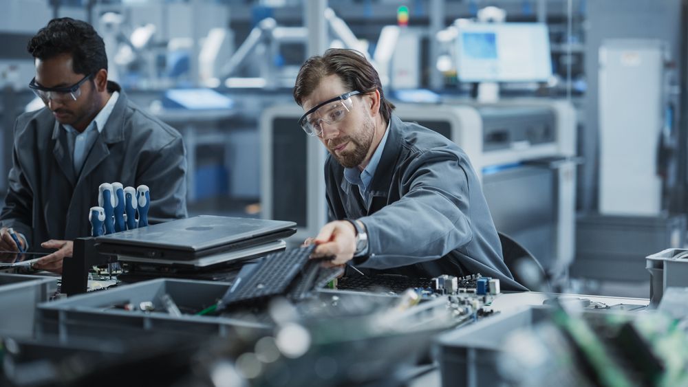 Indian And White Male Workers Taking Apart Laptops To Recycle For Production Of Sustainable Devices At Automated Electronics Factory With Robotic Arms.