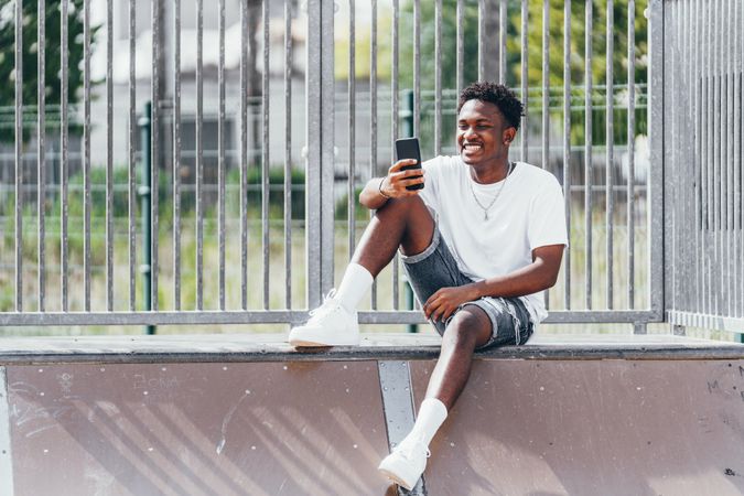 Sportive black men thoughtfully browsing smartphone while sitting on fence