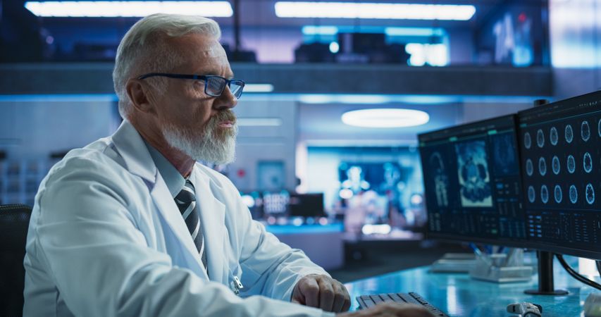 Male Neuroscientist Using Desktop Computer To Analyze MRI Scans Of Brain In Medical Research Center Laboratory.